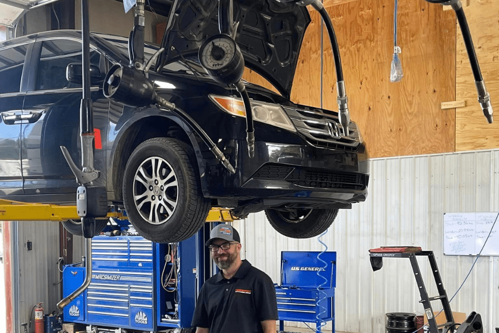 Full-Service Auto Repair in Jamestown, TN At Independent Auto and Diesel Repair. A mechanic smiles while standing in an auto repair shop under a black Honda minivan raised on a yellow vehicle lift, surrounded by hanging service hoses and blue toolboxes.